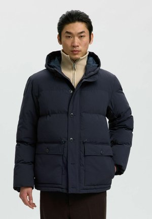 Homme portant une doudoune bleu marine par-dessus un pull beige à zip, debout devant un fond gris clair uni.