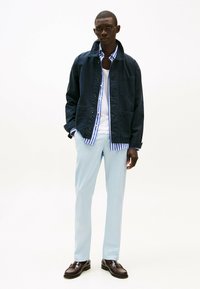 Chaqueta de lona azul marino con cierre de botón, dos bolsillos frontales; superpuesta a una camisa de rayas azul y blanca, combinada con pantalones ligeros azules y zapatos marrones.