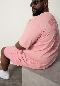 Růžový bavlněný set trička a šortek s grafickým potiskem na zádech. Model sedí na betonovém povrchu, má na sobě sluneční brýle a bílé tenisky.