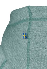 Ljusgrön tröja med strukturerad tyg, med kontrasterande teal-sömmar och en liten svensk flaggetikett på sidan.