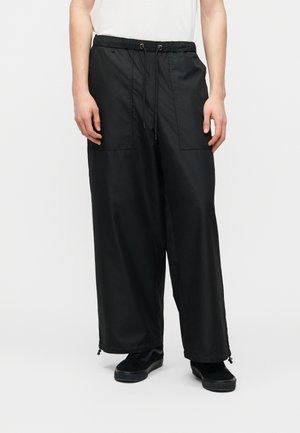 Personne portant un pantalon ample noir à cordon de serrage avec poches plaquées, associé à des baskets noires et un haut blanc, debout devant un fond uni.
