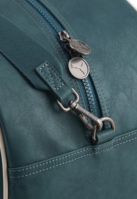 Afdeling af en turkis lædertaske med en stofrem, metal lynlås træk med Puma-logo og en metallukning graveret med "PUMA".