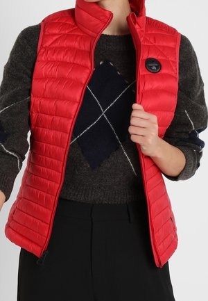 Personne portant un gilet matelassé rouge par-dessus un pull gris foncé avec un grand motif losange noir et blanc, tenant le col du gilet.