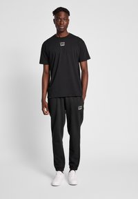 Sort bomulds T-shirt med rund hals og logo, parret med sorte fleecebukser med elastiske manchetter og logo, komplementeret af hvide sneakers.