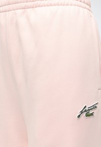 Tissu rose clair avec taille élastique, logo crocodile vert brodé et inscription noire en bas à droite.