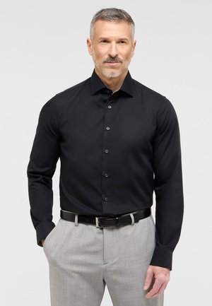 Schwarze Langarm-Hemd mit Knopfverschluss, kombiniert mit hellgrauen Hosen und einem schwarzen Leder-Gürtel. Glatte Stofftextur.
