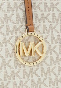 Eticheta de geantă Michael Kors prezintă un logo circular auriu cu inițialele "MK", atașată printr-o curea maro pe un fundal crem cu model.