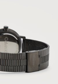 Bracelet et boucle de montre Calvin Klein en métal noir avec gravure de la marque, montrant partiellement le dos de la montre sur un fond blanc.