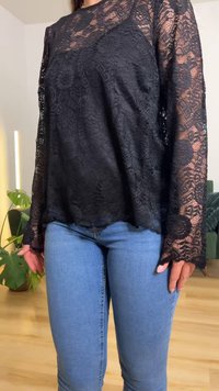 Vrouw draagt een zwart kanten top met lange mouwen en lichtblauwe strakke spijkerbroek, staand binnen bij een groene plant en een bank.