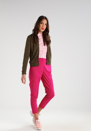 Femme debout portant une veste vert olive, une chemise rose à boutons, un pantalon rose vif fuselé et des baskets rose clair sur un fond uni.