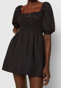 Nainen, jolla on päällään musta lyhyt mekko, jossa on puffihihat, rypytetty yläosa, neliönmuotoinen kaula-aukko ja hopeaketju kaulassa.