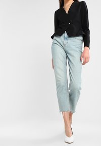 Kvinna i en svart blazer, ljusblå jeans med fransar och vita spetsiga klackskor mot en enkel vit bakgrund.