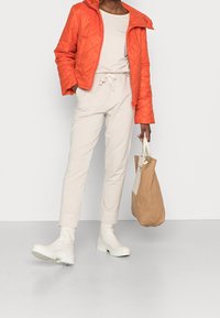 Giacca arancione trapuntata, top beige, joggers beige con vita regolabile, stivaletti bianchi alla caviglia, che tiene una borsa tote marrone. Texture morbide, look moderno.