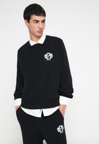 Czarny prążkowany sweter z okrągłym dekoltem, ozdobiony białym haftowanym logo, noszony na białej koszuli z kołnierzykiem. Dopasowane czarne spodnie z detalem logo.