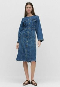 Denim-kjole med lange ermer, knappestolpe foran, to brystlommer, midtlengde, blå farge, og en tettsittende midje. Brukt med svarte og brune leppestifter.