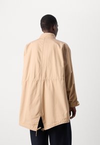 Beige lett jakke med lange ermer, bakbelter og snorjustering nederst. Har stående krage og knappeledd.