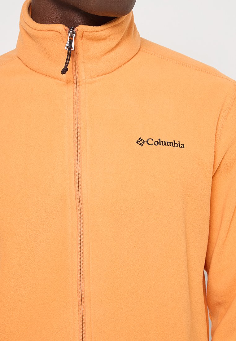 Zbliżenie na osobę w brzoskwiniowej kurtce zamkowej z logo Columbia na piersi, ukazujące wysoki kołnierz i suwak.