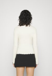 Person med lockigt mörkt hår som har på sig en figurnära vit långärmad topp och en svart minikjol, stående vänd bort från kameran mot en vit bakgrund.