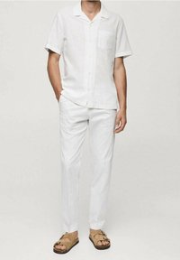 Camicia di lino bianca a maniche corte con tasca, abbinata a pantaloni bianchi e sandali marroni, caratterizzata da una vestibilità rilassata e una texture liscia.