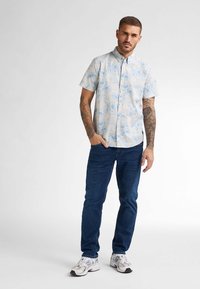 Hemdkorte mouwen in lichtblauw met bloemenpatroon, gecombineerd met donkerblauwe jeans en witte sneakers. Kenmerkende button-down kraag.