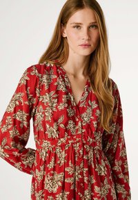 Robe florale rouge avec un décolleté en V, des manches longues, une taille cintrée et des motifs floraux beiges sur un tissu doux. Détails de boutons simples visibles.