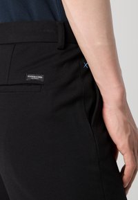 Czarne spodnie wykonane z miękkiego materiału, z tylną kieszenią z metką z logo i detalem jasnoniebieskiego przeszycia. Krój slim fit.