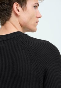 Svart rippad tröja med rund halsringning och texturerat mönster, med sömdetaljer längs axlarna och ryggen.