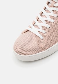 Gros plan d'une basket tricotée rose clair avec des lacets blancs et une semelle en caoutchouc blanche sur un fond clair.