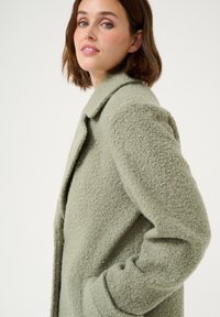 Groene, textuurrijke wollen jas met een kraagontwerp, opgestikte zakken en een ontspannen pasvorm, die een vlezige oppervlakte en een verfijnde structuur laat zien.