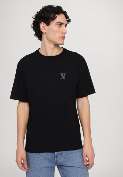 Black cotton t-shirt with short sleeves, round neckline, and a small embroidered logo on the left chest. Paired with light blue jeans.