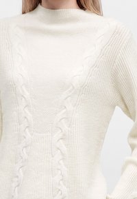 Femme portant un pull en maille couleur crème avec un col montant et des motifs torsadés en relief sur le devant et les manches.