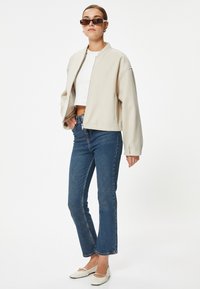 Beige kort bomberjacka med rund krage, bärs över en vit topp, ihop med blå utsvängda jeans och vita ballerinas.