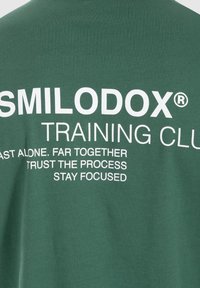 Camiseta de algodón verde con texto impreso en blanco en la parte trasera. El texto incluye el nombre de la marca y frases motivacionales. Diseño de manga corta.