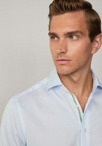 Camicia button-up azzurro chiaro con colletto alla francese, caratterizzata da dettagli verdi lungo la patta. Tessuto liscio con una texture sottile.