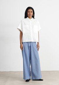White short-sleeve button-up shirt with two front pockets, paired with loose light blue wide-leg trousers. Smooth fabric, minimal design.