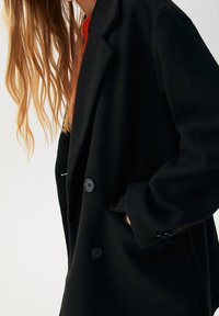 Cappotto lungo nero in lana con colletto a punte, due bottoni e tasche laterali. Il tessuto ha una superficie liscia e una vestibilità comoda.