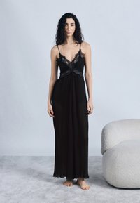 Γυναίκα με μακριά, σγουρά μαλλιά στέκεται ξυπόλυτη σε χαλί φορώντας μαύρο φόρεμα με δαντέλα δίπλα σε μια λευκή μοντέρνα καμπυλωτή πολυθρόνα.