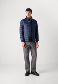 Young man wearing a navy quilted jacket, gray trousers, black sneakers, standing against a plain white background.
