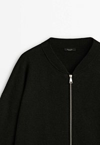 Pull noir à fermeture éclair avec col côtelé et fermeture éclair argentée, fabriqué en tissu tricoté doux, présenté de la partie supérieure du torse sur un fond blanc.