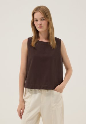 Mujer con cabello rubio hasta los hombros que lleva una camiseta sin mangas de color marrón oscuro y pantalones beige claro con la mano derecha en el bolsillo.