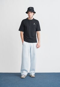 Svart bomulls-T-skjorte med logo, lyseblå joggebukser, svart bøttehatt med logo og beige joggesko med svarte detaljer på blått gulv.