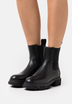 Bottes noires en cuir pour la cheville avec des semelles épaisses portées sur des jambes nues sur fond blanc.