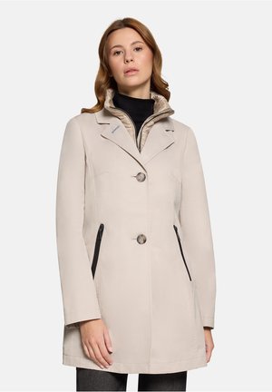 Kobieta nosząca beżowy płaszcz do połowy długości z dwoma guzikami i czarnymi zasuwanymi bocznymi kieszeniami, nałożony na beżową pikowaną kurtkę.