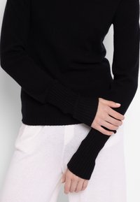 Prilegajoč črn pulover iz mehkega pletenega materiala, z rebrastimi manšetami in okroglim izrezom. Kombiniran z lahnimi hrapavimi hlačami.