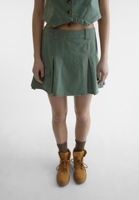 Grønn kort topp med knapper, matchende grønn plissert skjørt, og tan fargede snøresko med brune strømper. Glatt stoff, bruksvennlig design.