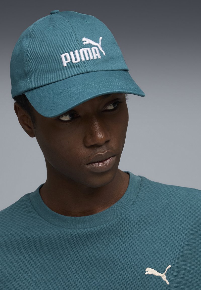 Homme portant une casquette Puma turquoise et un t-shirt texturé turquoise assorti avec le logo Puma blanc, regardant légèrement sur le côté sur fond gris.
