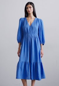Blauwe katoenen midi-jurk met een V-hals, pofmouwen en een geplooide taille. Beschikt over een gelaagde rok met een zachte, gestructureerde afwerking.