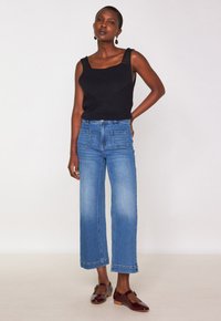 Donna che indossa un top nero senza maniche in maglia, jeans a gamba larga blu accorciati con tasche frontali, e scarpe Mary Jane con tacco basso di colore rosso scuro.