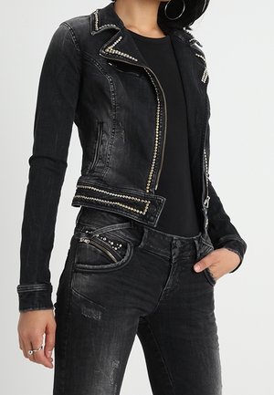 Femme portant une veste en jean noire cintrée et un jean noir avec des détails cloutés et une fermeture éclair, main dans la poche, sur un fond clair uni.