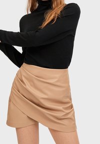Suéter negro de cuello alto combinado con una falda envolvente de imitación cuero beige, que presenta un diseño arrugado y una textura suave.
